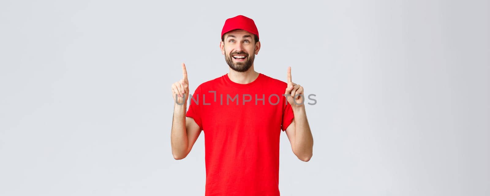 Online shopping, delivery during quarantine and takeaway concept. Cheerful excited courier in red uniform cap and t-shirt, smiling amazed and pointing fingers up, reading banner or sign by Benzoix