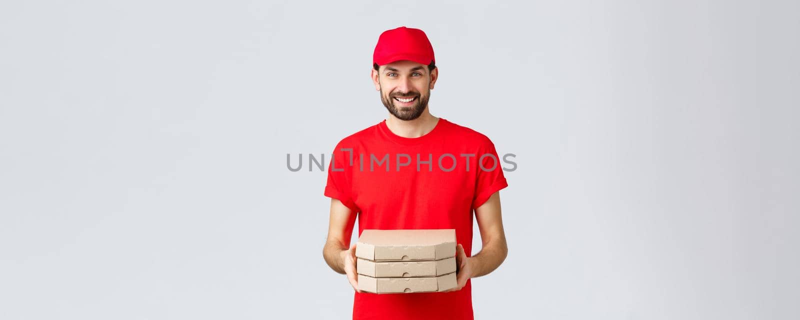 Food delivery, quarantine, stay home and order online concept. Friendly smiling courier in red uniform giving client order. Employee bring pizza at your house, standing grey background by Benzoix