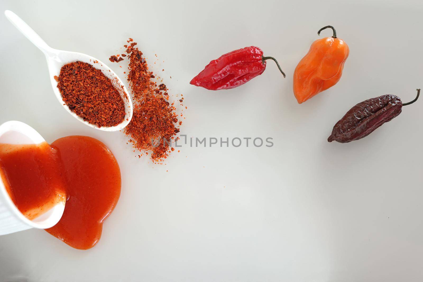 Three varieties of hot peppers Scotch bonnet, naga morich, bhut jolokiaand their derivatives - sauce and ground hot pepper on a white glass surface