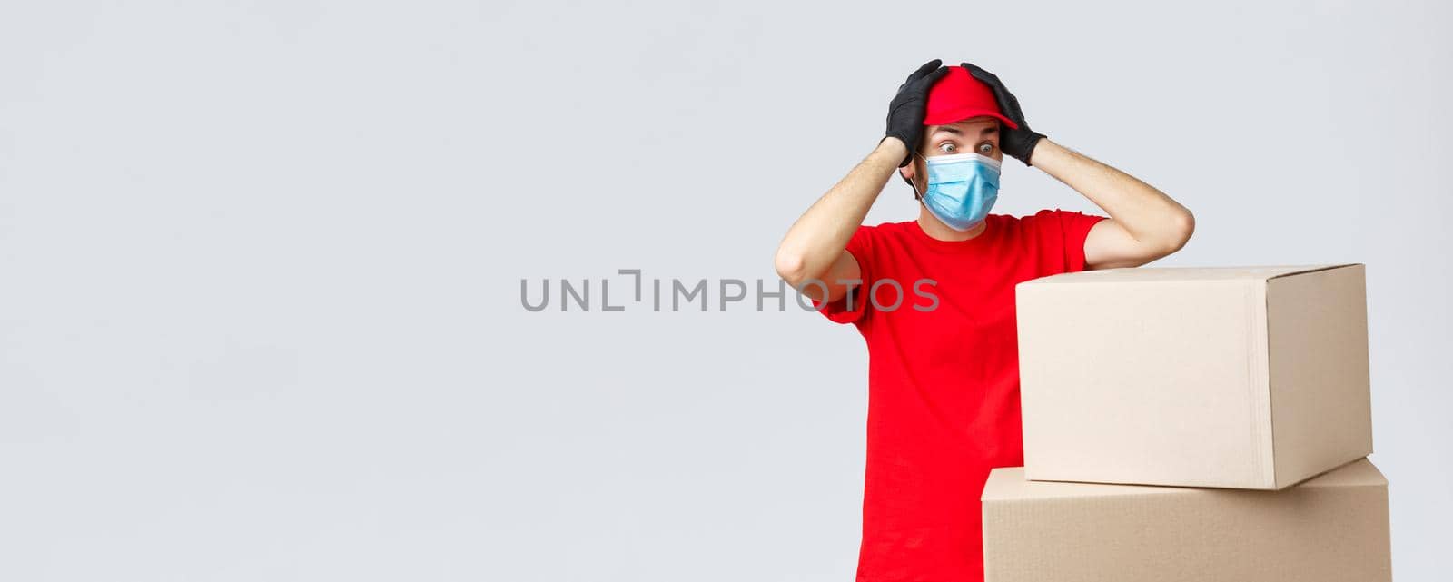 Packages and parcels delivery, covid-19 quarantine and transfer orders. Concerned and troubled courier in red uniform, face mask and gloves, grab head and gasping shocked staring at boxes by Benzoix