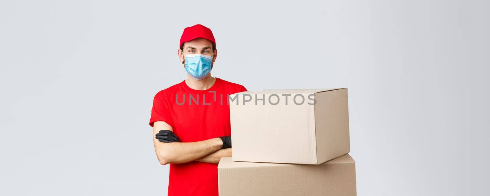 Packages and parcels delivery, covid-19 quarantine and transfer orders. Confident young courier in red uniform, gloves and medical mask, cross arms as standing boxes, grey background.