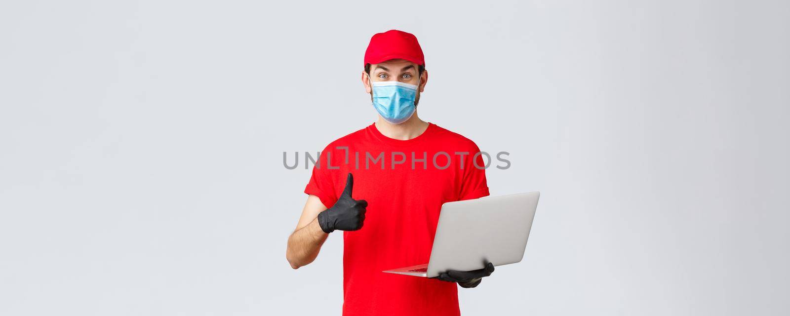 Customer support, covid-19 delivery packages, online orders processing concept. Enthusiastic courier in red uniform thumbs-up reading promo on webpage. Delivery guy hold laptop, wear mask and gloves by Benzoix