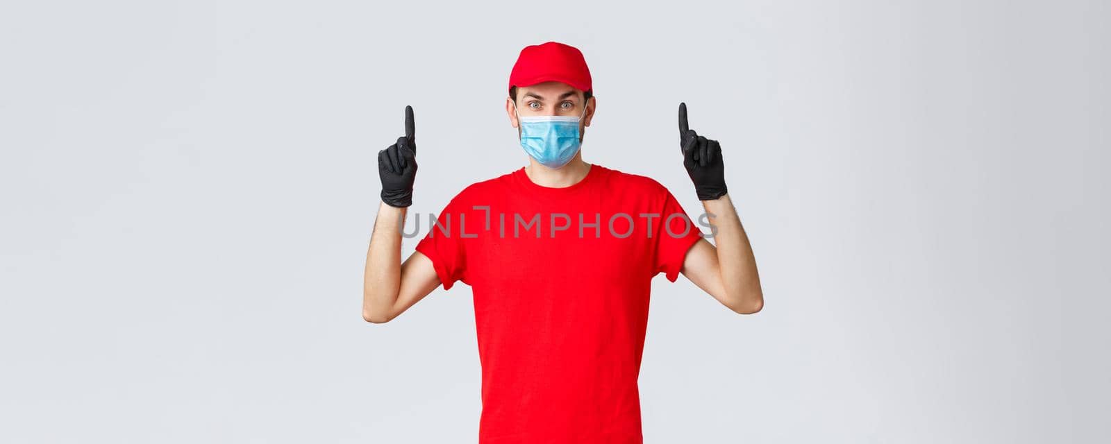 Covid-19, self-quarantine, online shopping and shipping concept. Surprised delivery guy in red uniform and cap, raising eyebrows amazed or excited, showing promo top, pointing up, wear medical mask by Benzoix