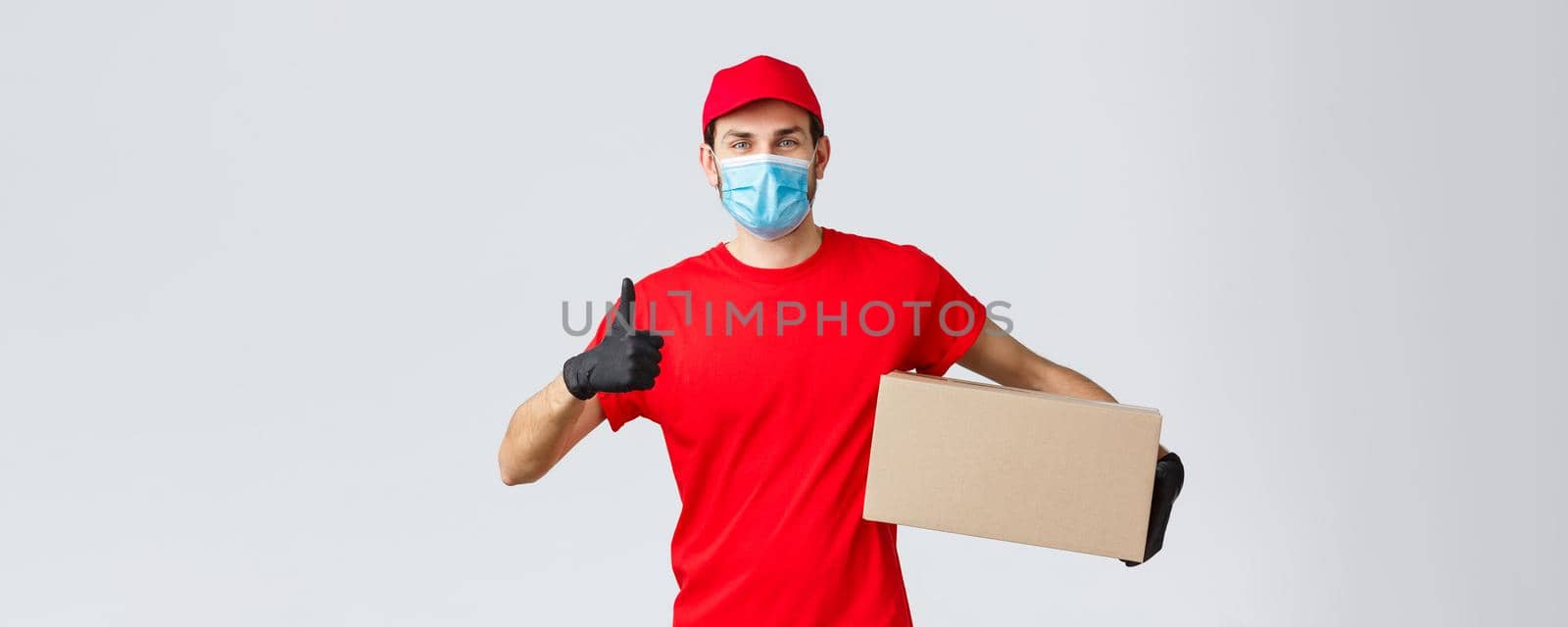 Packages and parcels delivery, covid-19 quarantine delivery, transfer orders. Cheerful courier in red uniform, gloves and face mask, thumb-up, recommend contactless deliver, holding box with order by Benzoix