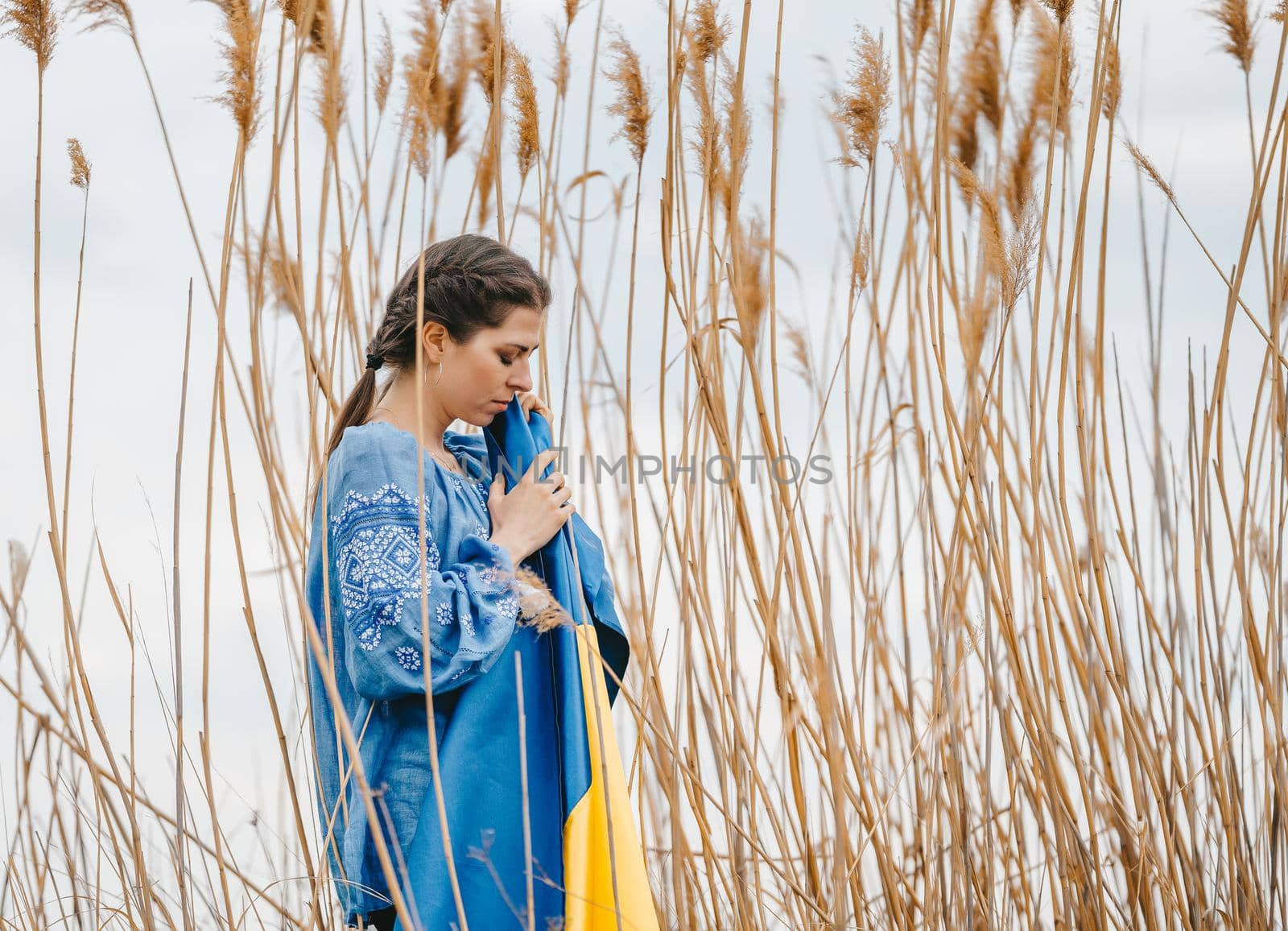 Sorrowful ukrainian woman with national flag on natural reeds background. Lady in blue embroidery vyshyvanka blouse. Ukraine, independence, freedom, patriot symbol, victory in war. High quality photo