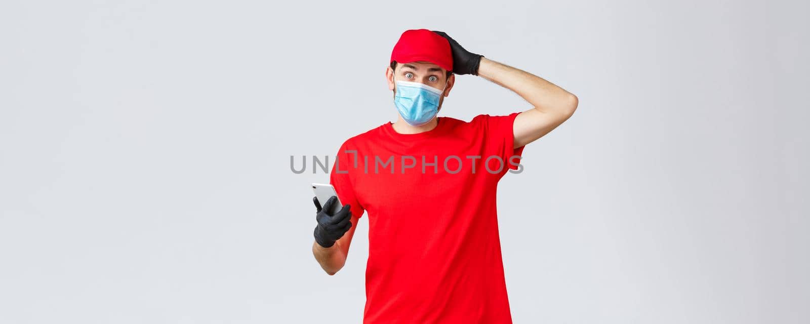 Contactless delivery, payment and online shopping during covid-19, self-quarantine. Confused and indecisive courier dont know, wear face mask and gloves, scratch head, hold smartphone by Benzoix