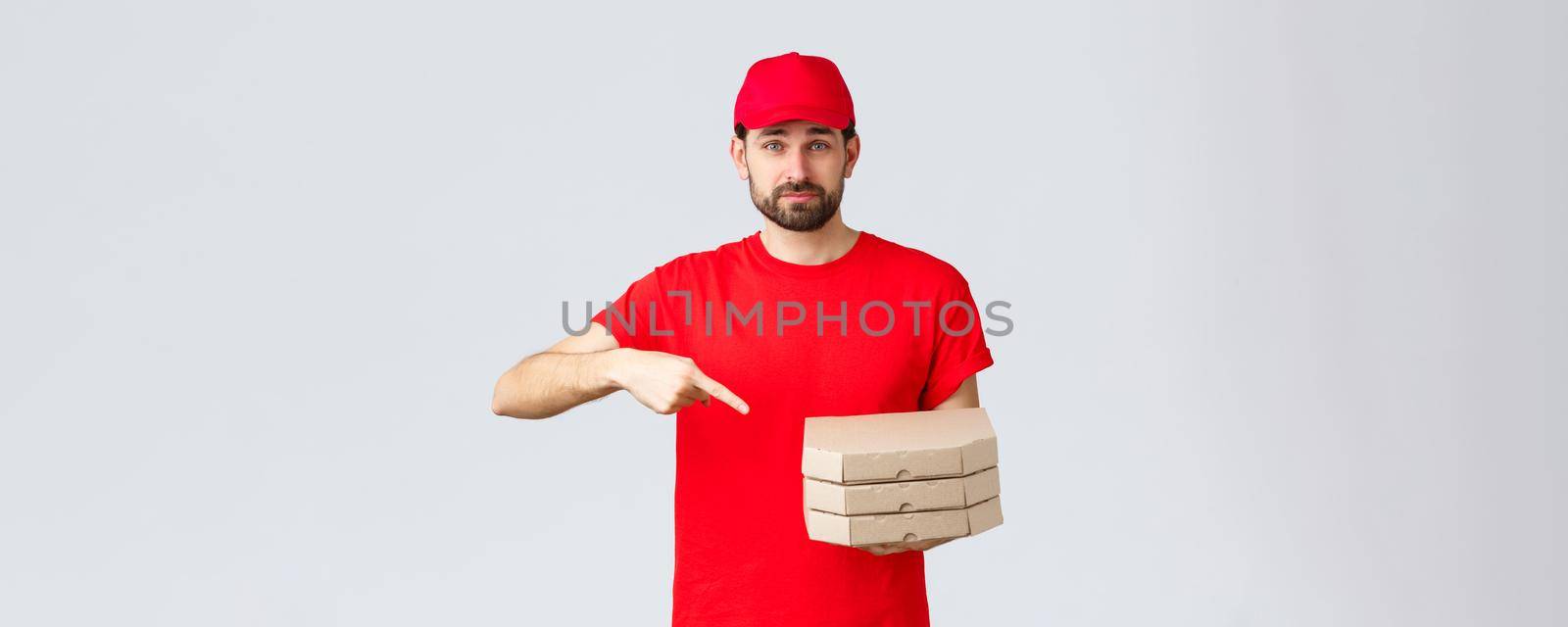 Food delivery, quarantine, stay home and order online concept. Confident friendly courier in red uniform cap and t-shirt, employee bring order pizza, pointing finger at boxes, grey background.