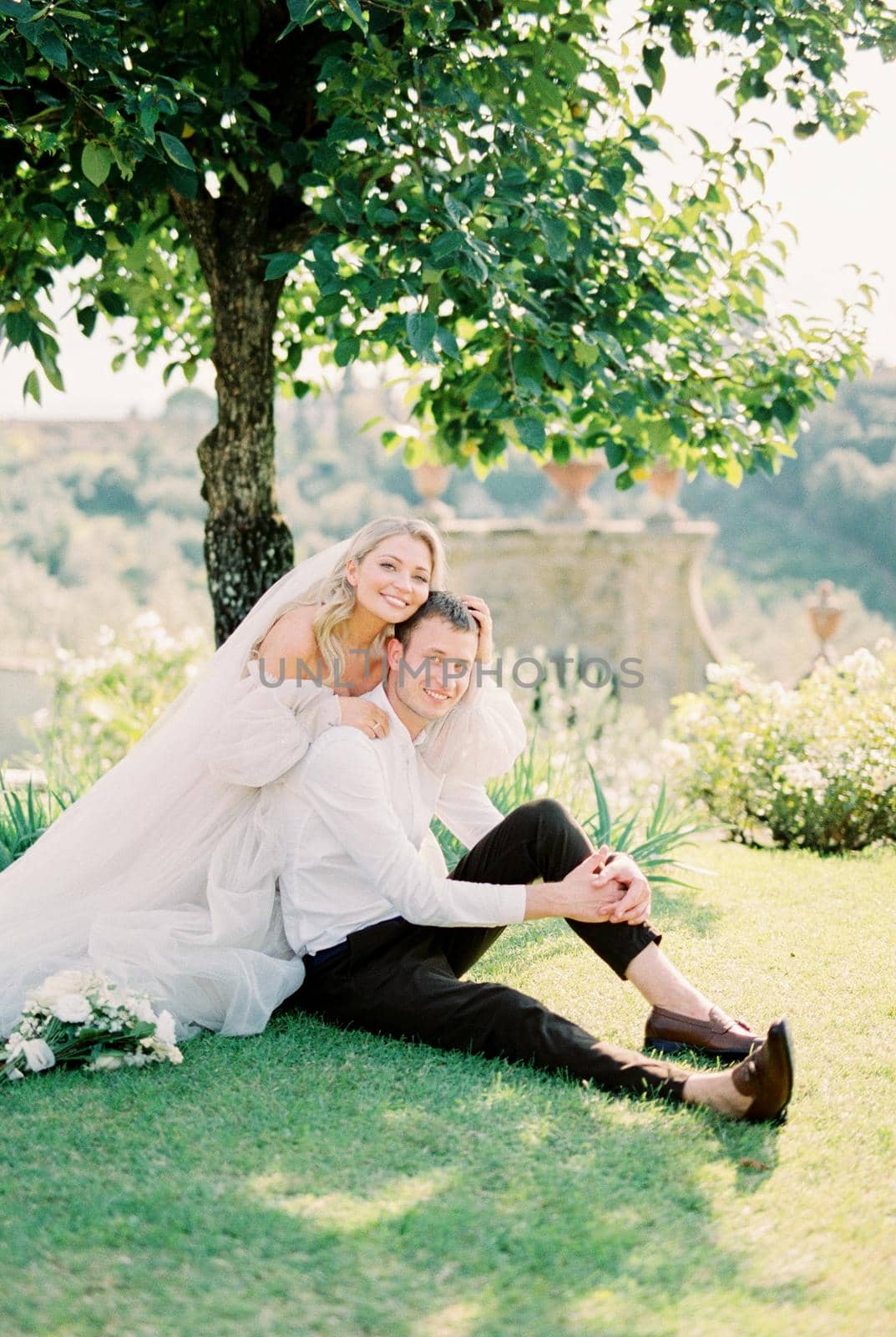 Bride hugs groom from behind, sitting under a tree in a green garden. Florence, Italy. High quality photo