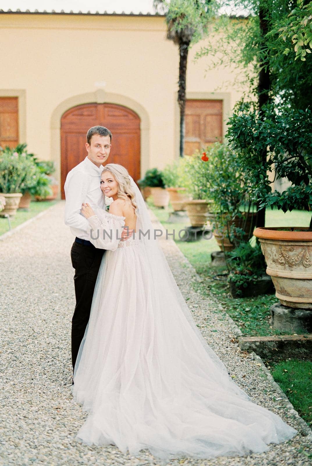 Bride and groom stand embracing on a gravel path near an old villa. High quality photo