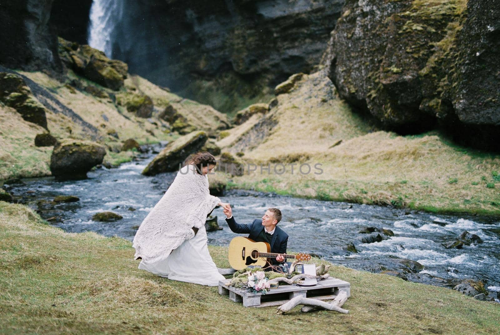 Groom sits with a guitar next to bride standing in a blanket on the river bank. High quality photo