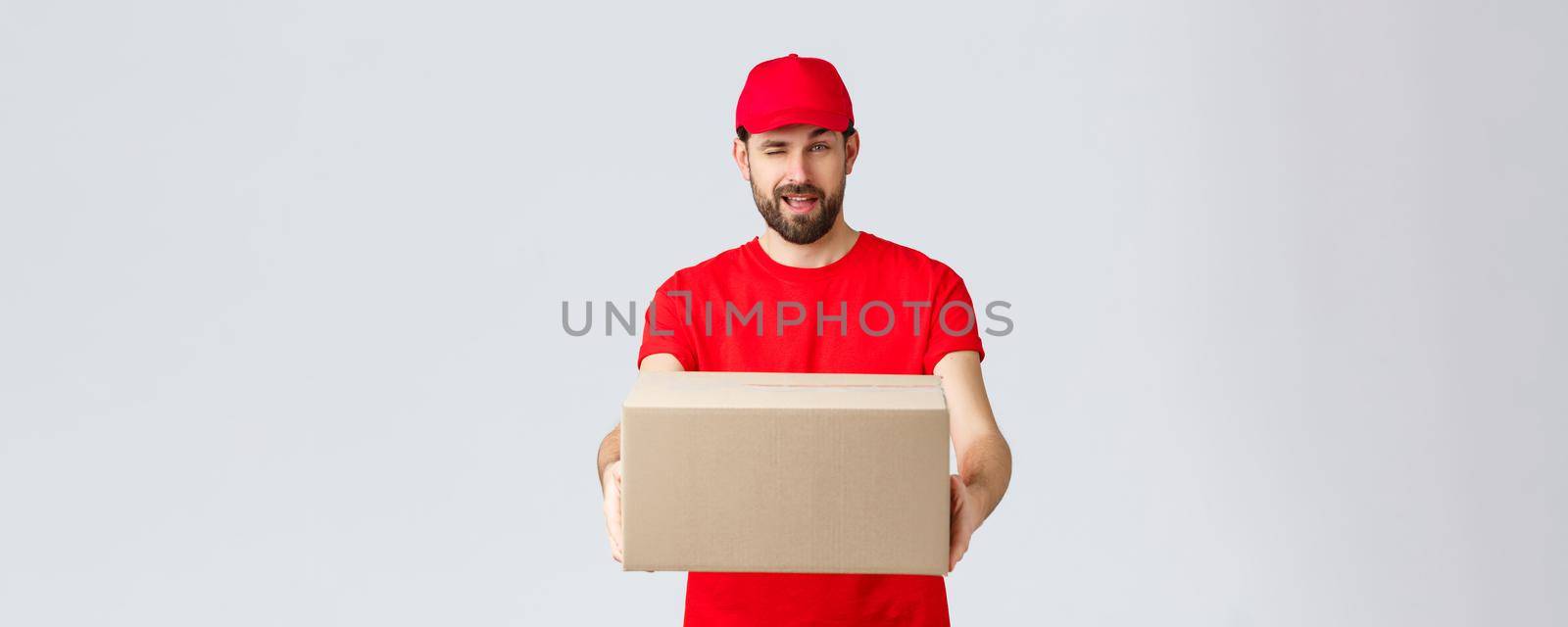 Order delivery, online shopping and package shipping concept. Cheeky handsome bearded courier in red uniform, handing box package to client. Employee wink to you and give order parcel.