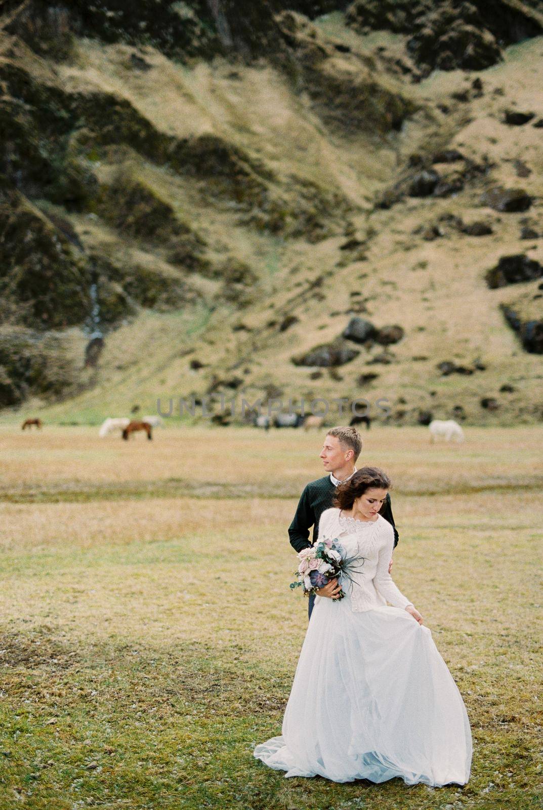 Bride with a bouquet stands next to her groom in a mountain valley. Iceland by Nadtochiy