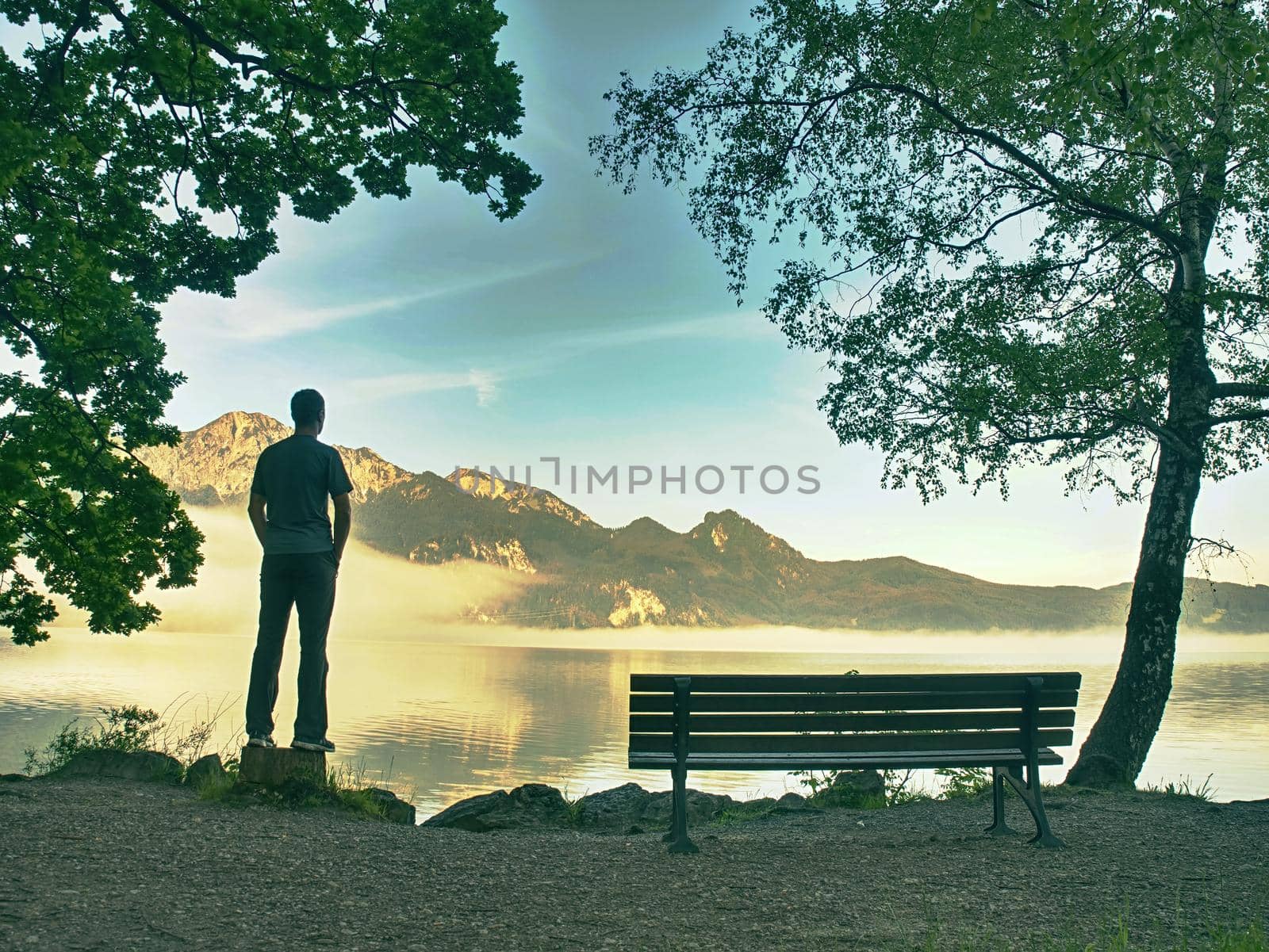 Alone man sits on bench beside an azure mountain lake. Man relax and watch high peaks of Alps above lake mirror.