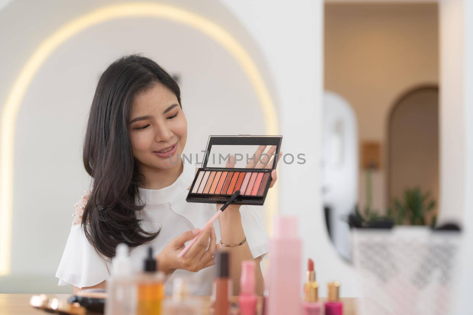 Asian female beauty blogger live streaming to review makeup product on social media, Modern young woman influencer demonstrating her daily cosmetic while talking on smartphone camera by nateemee