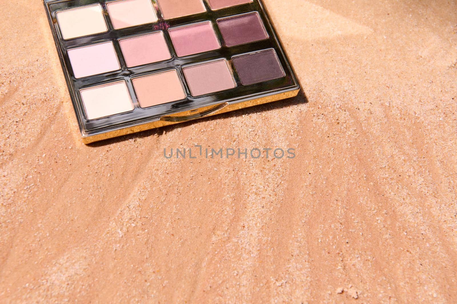 Partial overhead view of a makeup eyeshadows palette with shades of beige colors on a golden sandy background with copy ad space