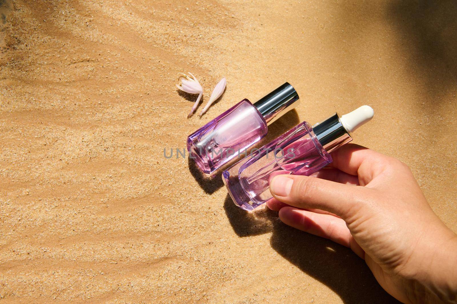 Close-up. Hand putting skin care beauty product, packaged in a light violet glass bottle on golden sand background. Advertising shot of nourishing, rejuvenating, anti-aging cosmetics. Still life art