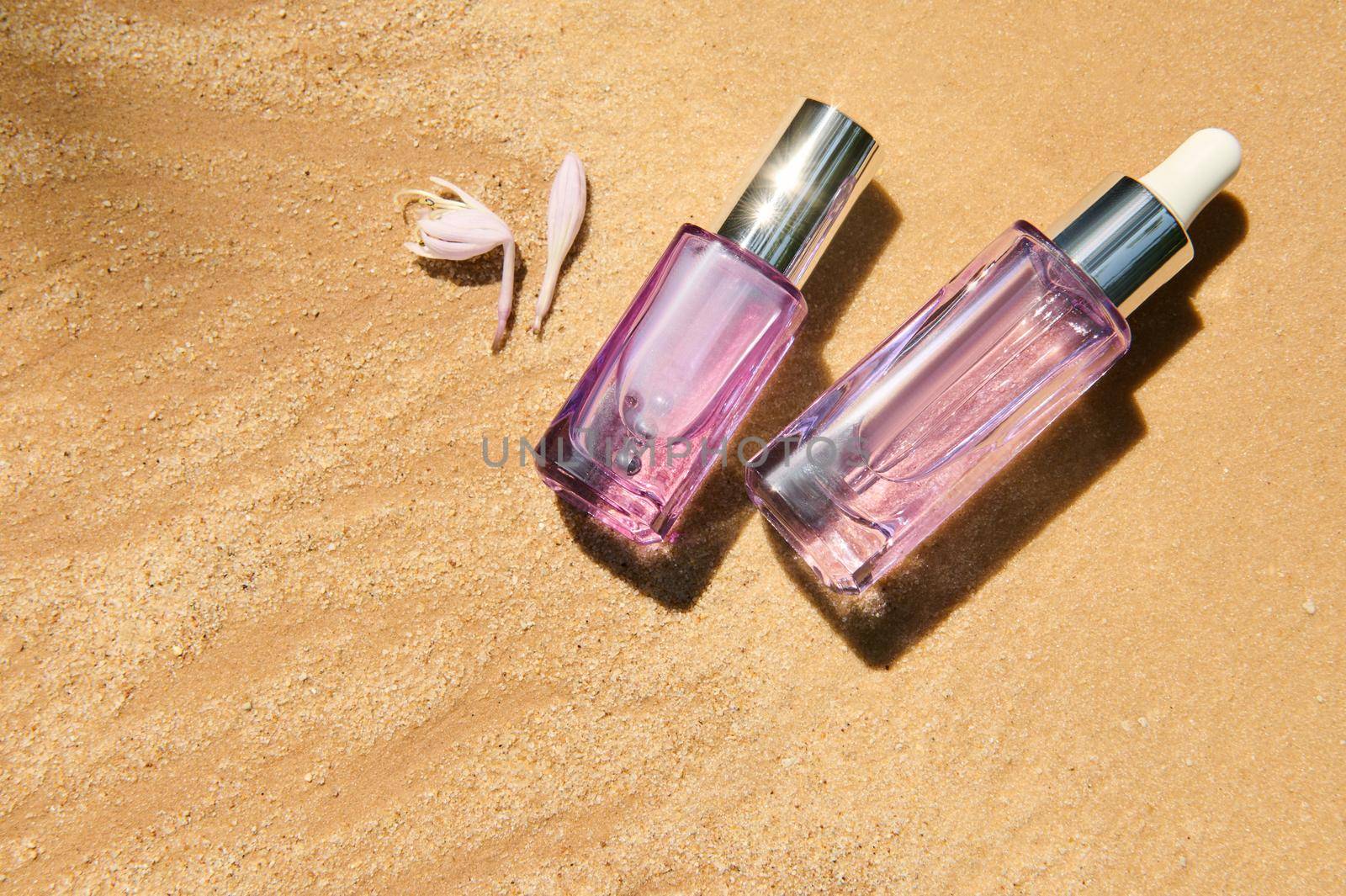 Flat lay of a liquid skin care product, packaging in a light violet glass bottles on sand background with sunlight and lily of the valley flower. Hyaluronic or polyglutamic acid oil cosmetic mockup.