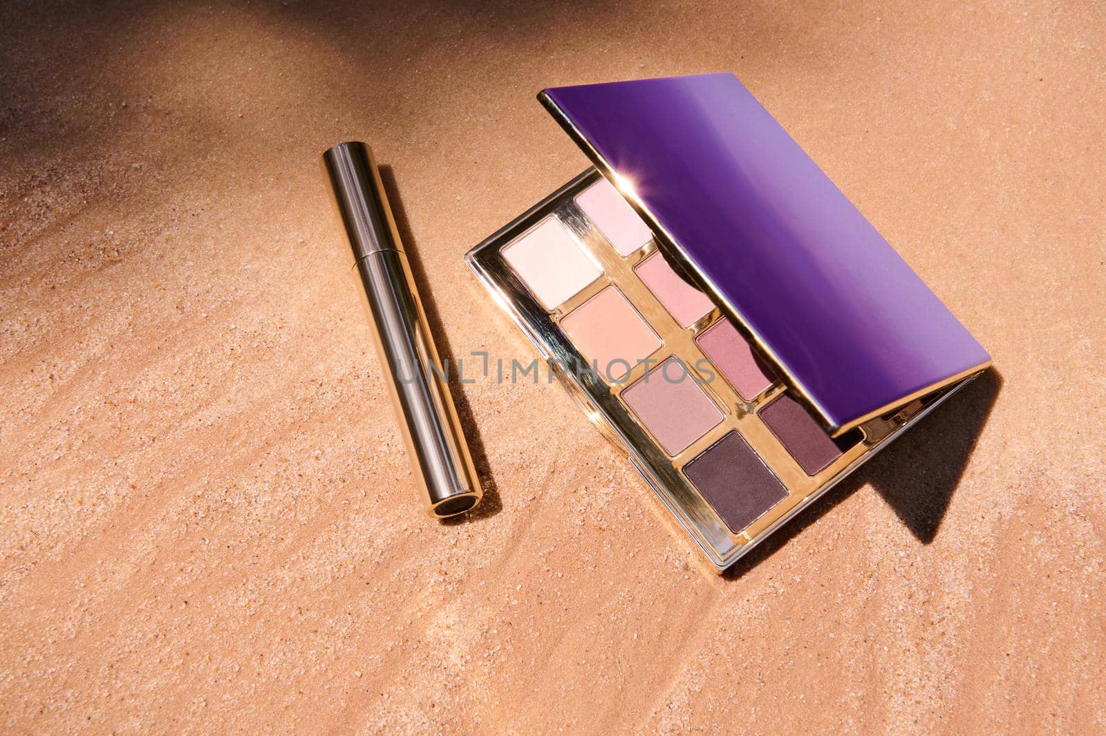 Top view. Glamour eyeshadow palette with gradient from dark brown to shades of beige and mascara on golden sand background with sunlight and shadow from palm leaves. Summer fashion. Makeup concept.