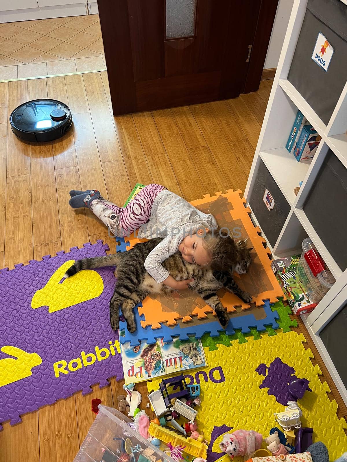 Little girl lies hugging a cat on a play mat by Nadtochiy