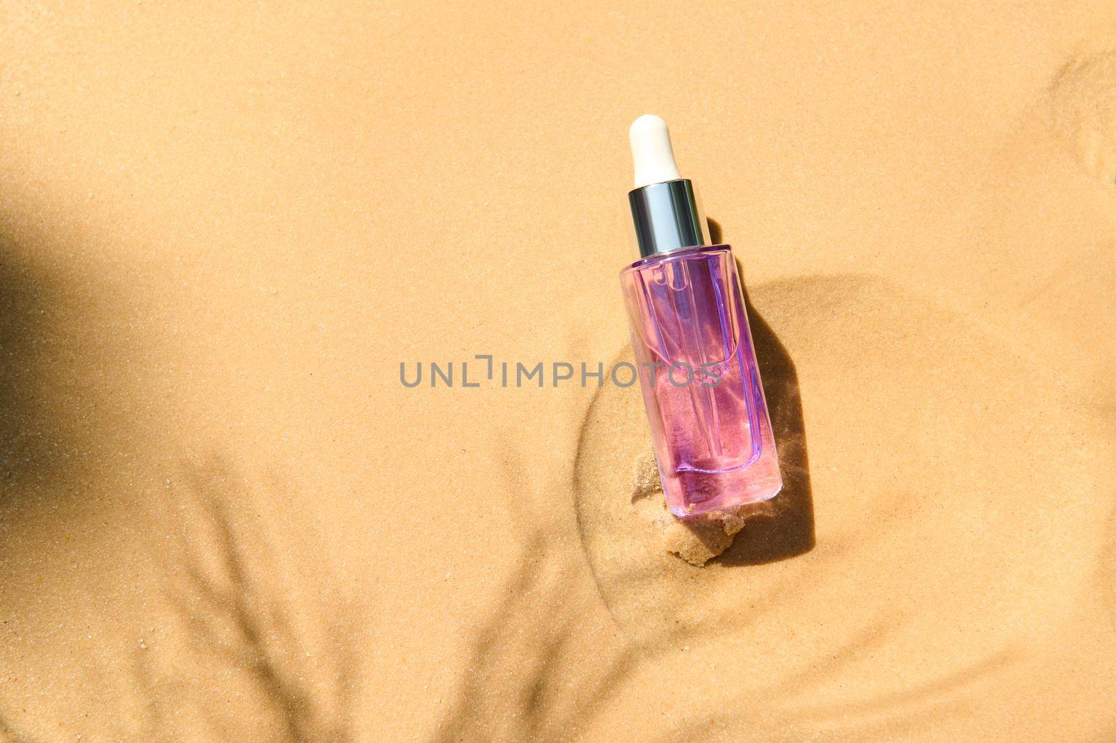 Anti aging serum with collagen and peptides in light violet glass bottle with dropper on sandy background with sunlight and shadow from palm leaf. Skincare essence for beautiful healthy skin. Top view