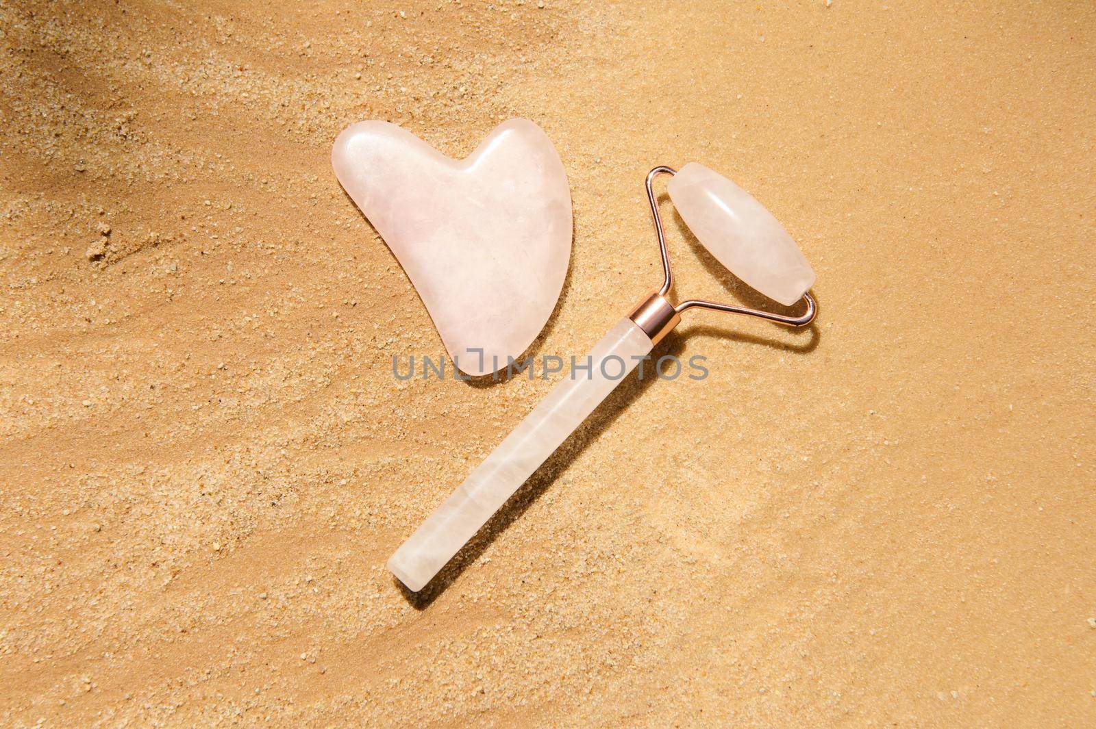 Flat lay. Minimalist still life art of a jade roller of pink stone and gua-sha massager, for home face lifting and lymphatic drainage massage, isolated on a golden sand background with copy ad space