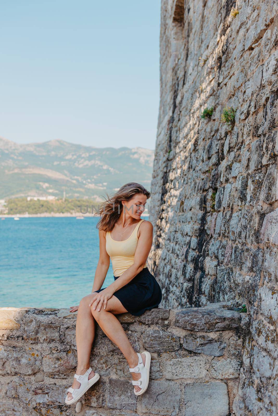 Beautiful girl sitting on a stone wall, in background is the blue sea, Budva, Montenegro