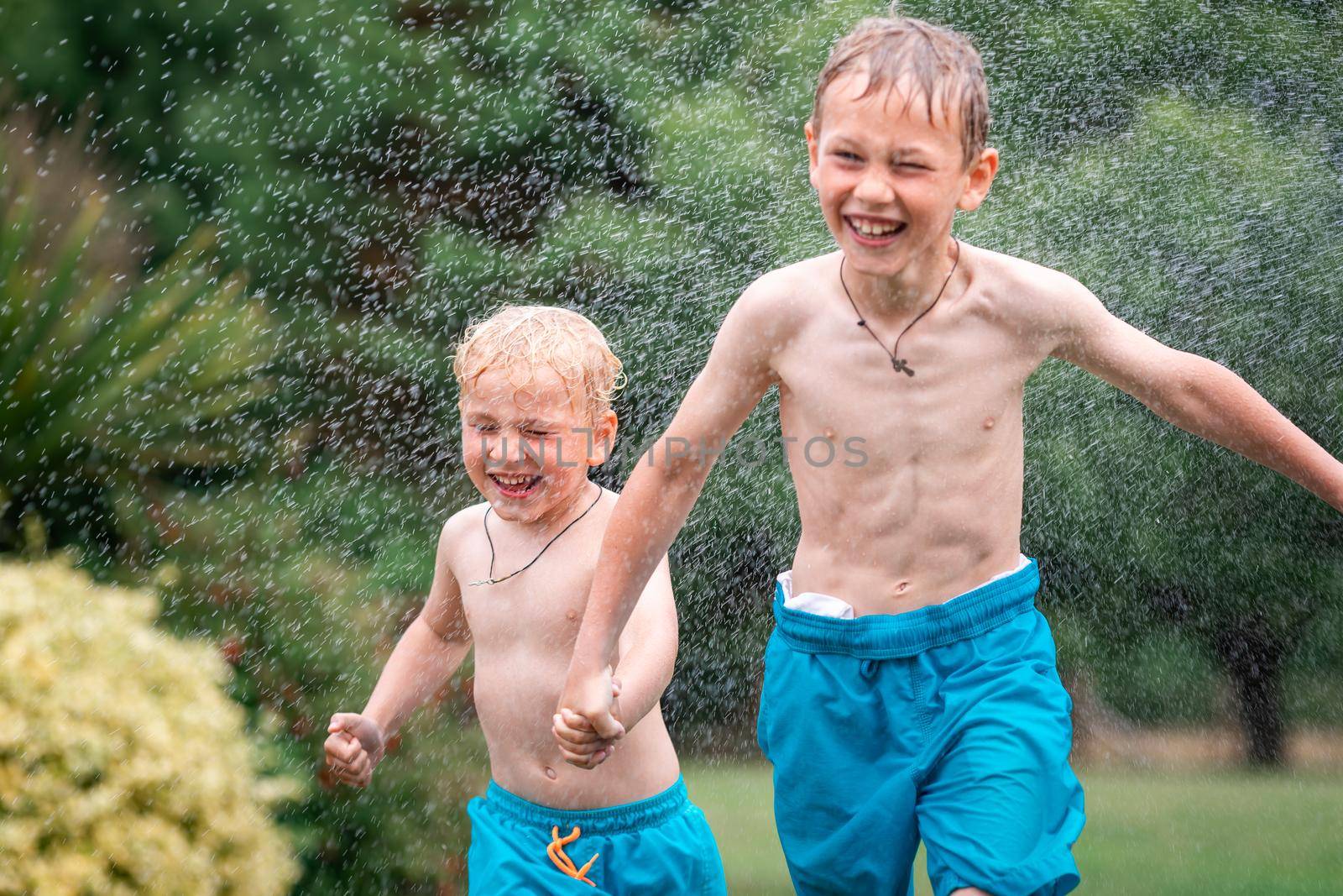 Kids play with water on hot summer day. Children with garden sprinkler having fun. Outdoor fun. Boys run on a field under water drops.