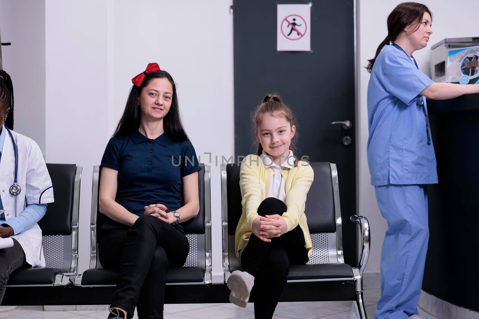 Smiling mother and daughter in busy private clinic waiting for pediatric doctor to do routine checkup by DCStudio