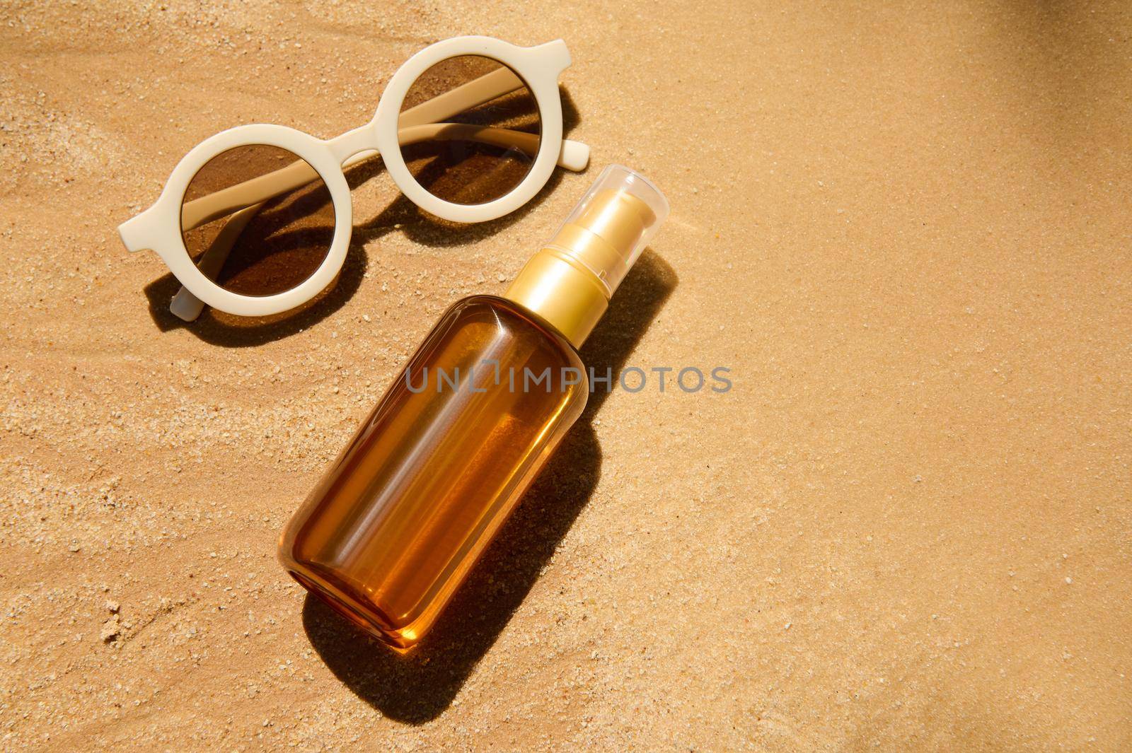 Top view. Minimalist still life of unbranded dark glass bottle with SPF oil or sunscreen body lotion, sunglasses on golden sand beach background. Summer cosmetic concept. Copy ad space for text