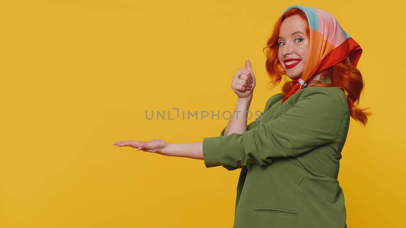 Beautiful redhead woman showing thumbs up, pointing empty place, advertising area for commercial text, copy space for goods promotion. Young ginger girl. Studio shot indoors on yellow wall background