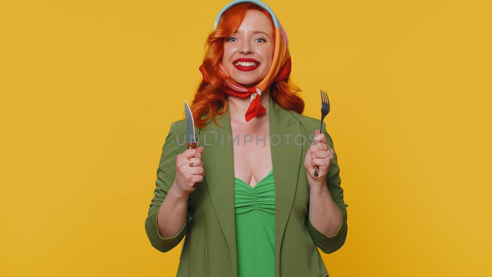 Ready to eat. Hungry redhead young woman in green jacket and dress waiting for serving dinner dishes with cutlery, will appreciate delicious restaurant meal. Ginger girl isolated on yellow background