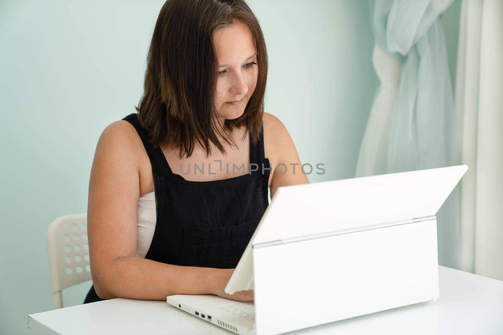 Girl is sitting in front of her white laptop transformer at desk in her house. Concept of remote work and education, side view, close-up