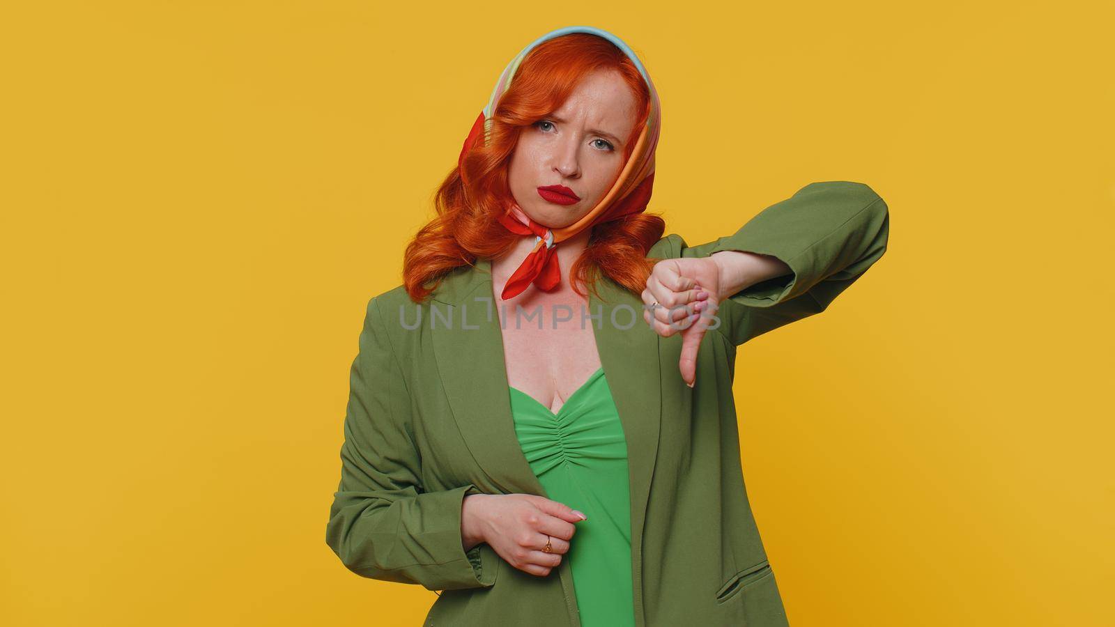 Upset unhappy red hair woman in jacket and kerchief showing thumbs down sign gesture, expressing discontent, disapproval, dissatisfied, dislike. Young ginger girl. Indoors studio on yellow background