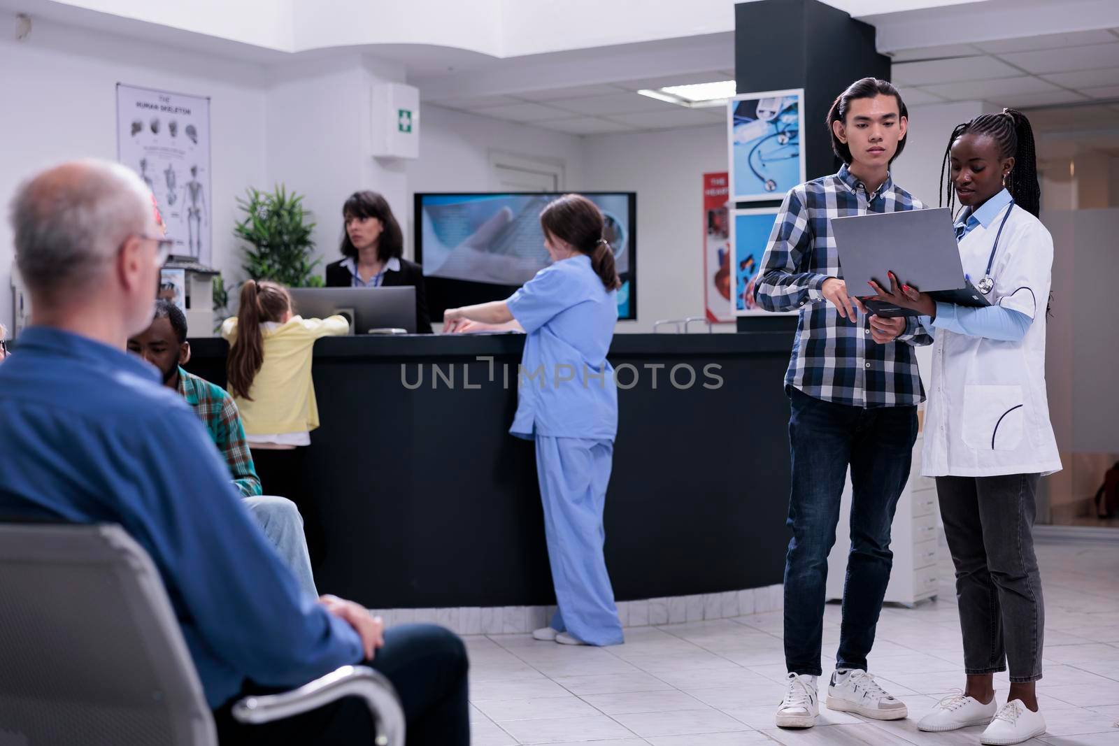 African american doctor holding laptop with mri scan results talking with asian patient in busy private clinic by DCStudio