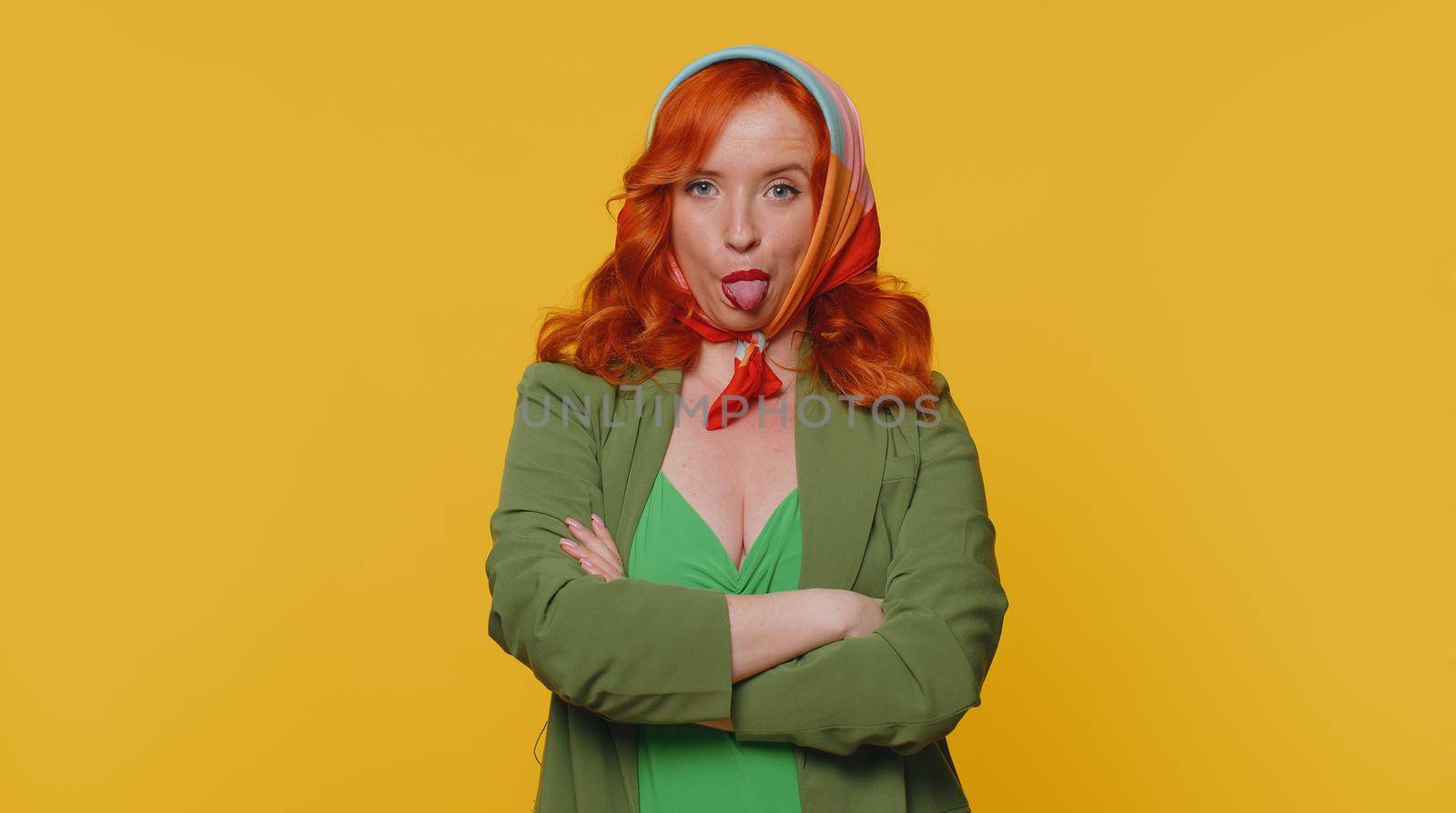 Funny joyful sincere redhead woman 20s in jacket making playful silly facial expressions and grimacing, fooling around showing tongue. Young beautiful ginger girl isolated alone on yellow background