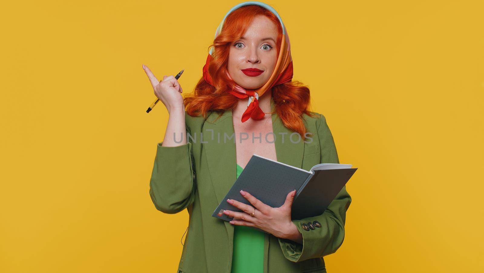 Thoughtful journalist redhead young woman making notes, writing down thoughts with pen into notepad notebook diary, to do list, good idea. Adult ginger girl indoor studio isolated on yellow background