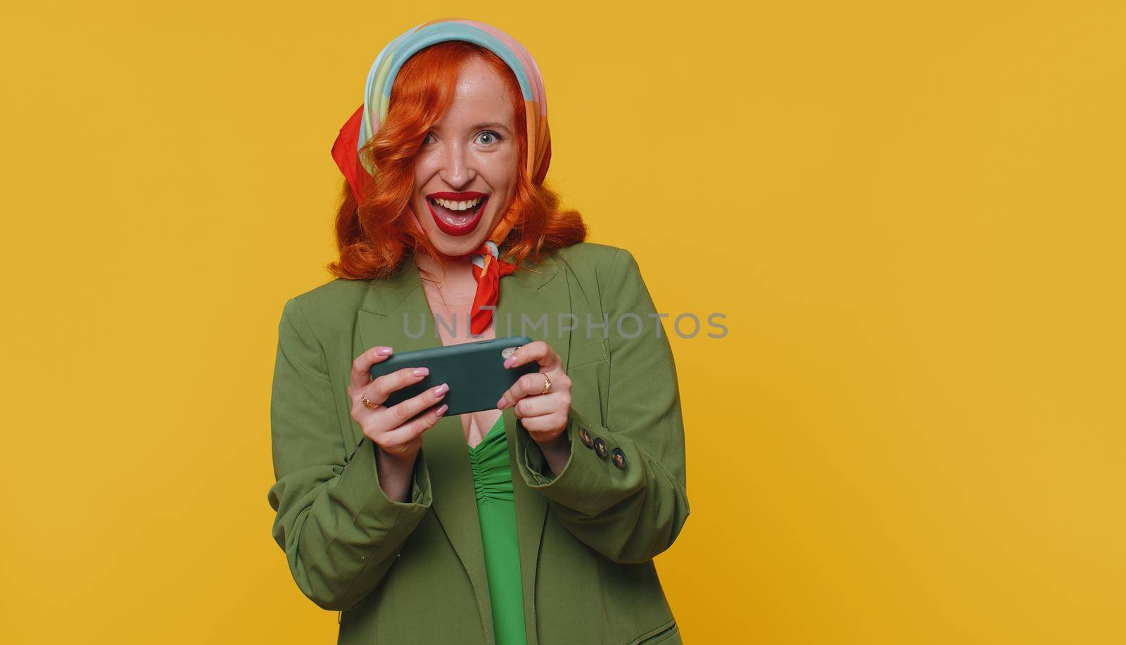 Worried funny addicted ginger woman enthusiastically playing racing video game on mobile phone. Young redhead girl using smartphone gadget app with drive simulator isolated on yellow studio background