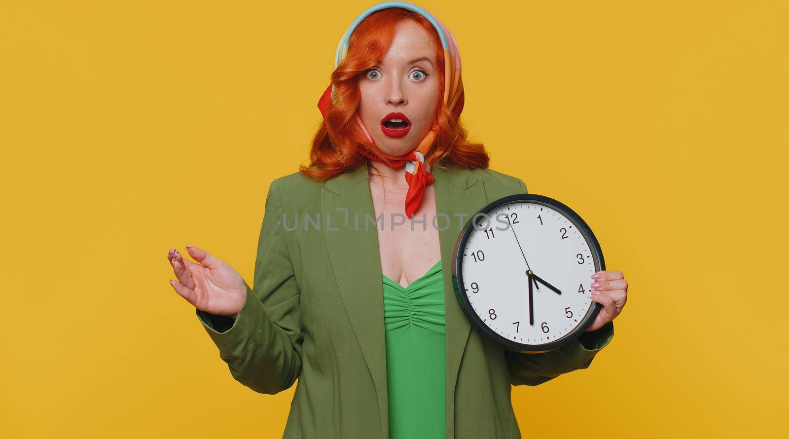 Redhead young woman in green jacket and dress with anxiety checking time on clock, running late to work, being in delay, deadline. Adult ginger girl indoors studio shot isolated on yellow background