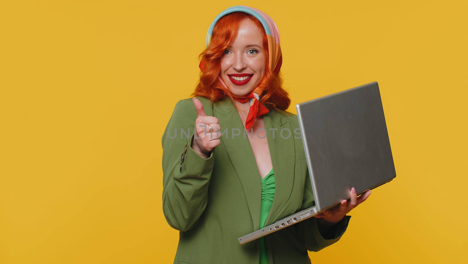 Redhead young woman working online on laptop finishing work, showing thumb up to camera, positive feedback, like, lottery win, approve good offer advertisement. Ginger girl on yellow studio background