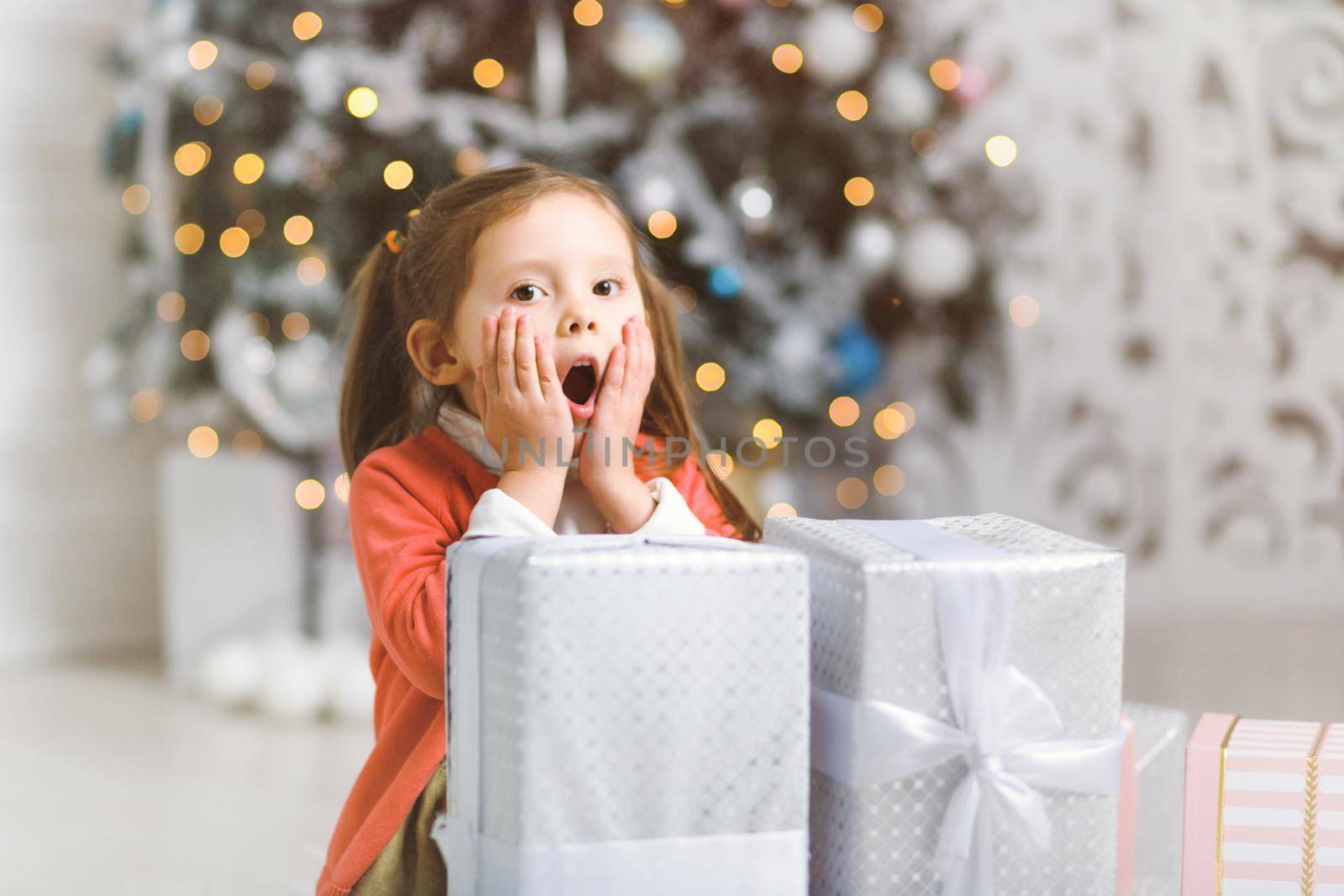 Cute little girl surprised by the abundance of Christmas gifts by a Christmas tree in cozy room.