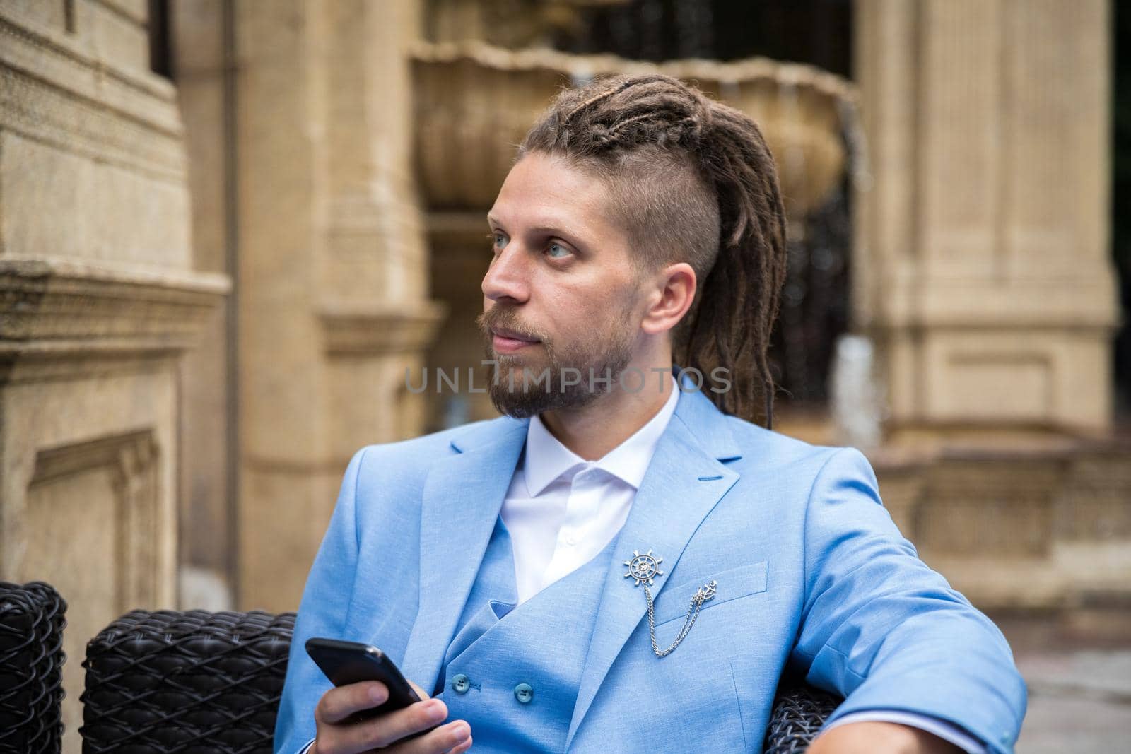 Young successful hipster caucasian man with dreadlocks in a blue suit with a smartphone in his hands sits in a street restaurant looks away.