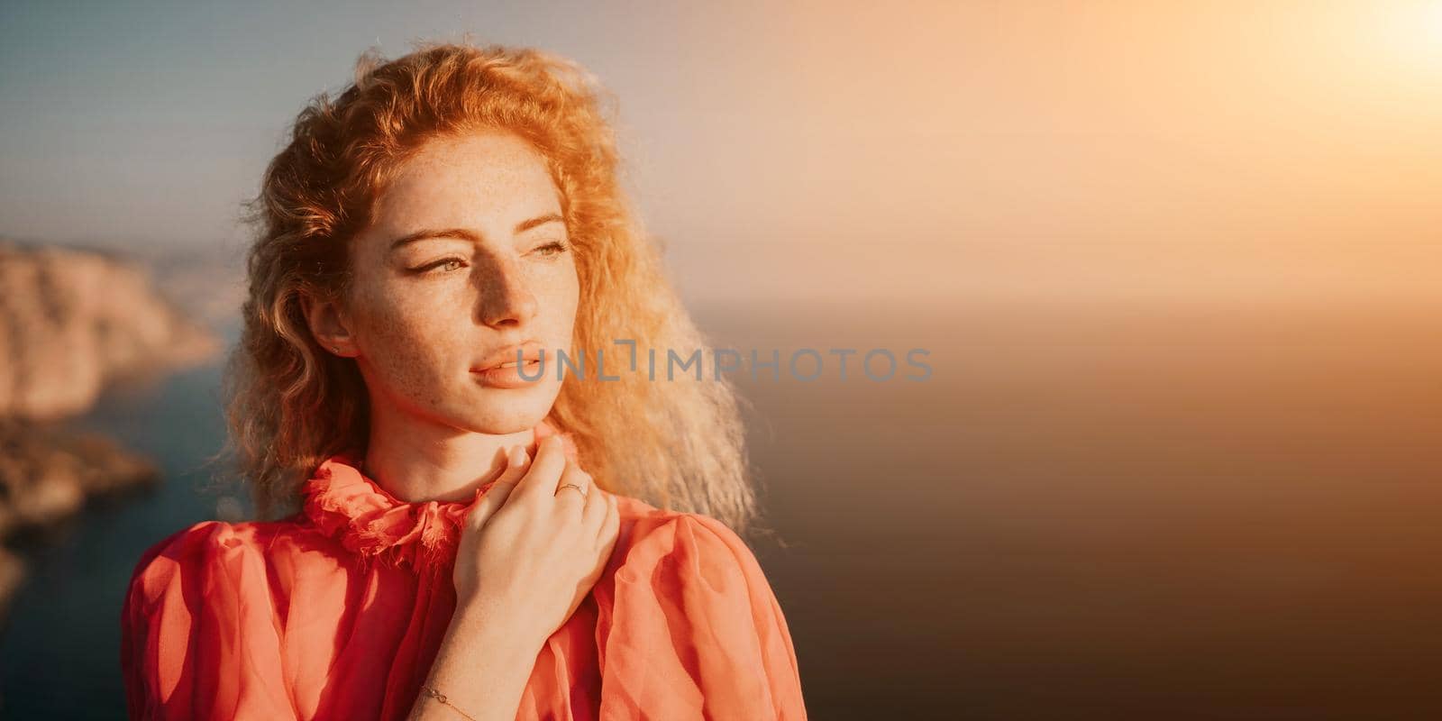 Close up shot of beautiful young caucasian woman with curly blond hair and freckles looking at camera and smiling. Cute woman portrait in a pink long dress posing on a volcanic rock high above the sea