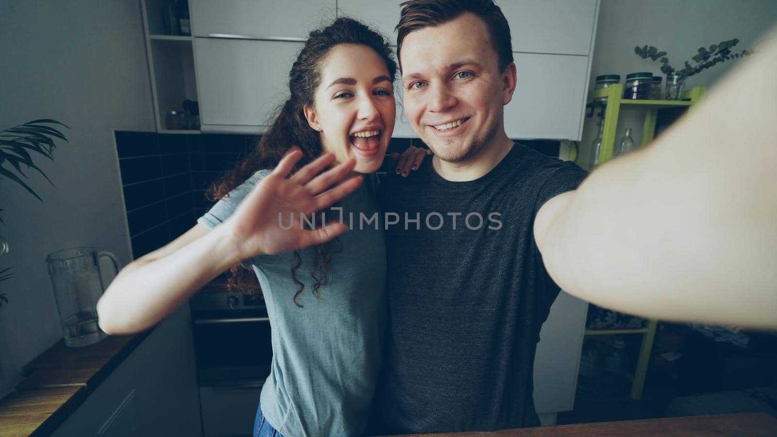 POV of Young happy couple having online video call with smartphone camera while standing in the kitchen at home by silverkblack