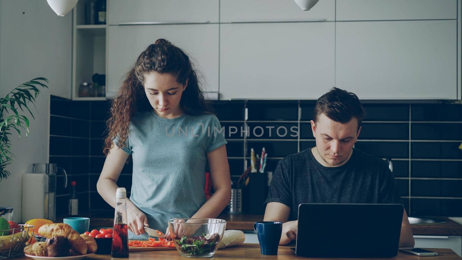 young caucasian couple at table, man is working on laptop, he is concentrated and pencive, woman is cooking cutting vegetables for salad, she is seriuos by silverkblack