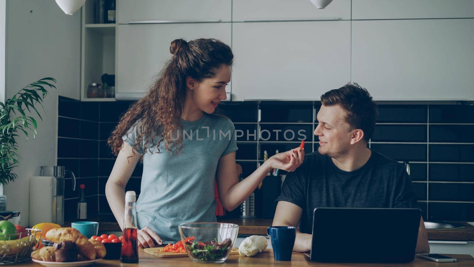 Cheerful couple at table woman cooking while gives hasband to try red pepper, man sitting working on laptop by silverkblack