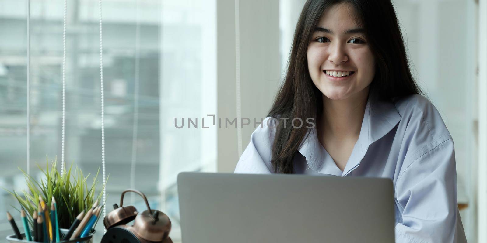 Young attractive happy asian female student sitting at living room floor smiling and looking up at camera on laptop at home by wichayada
