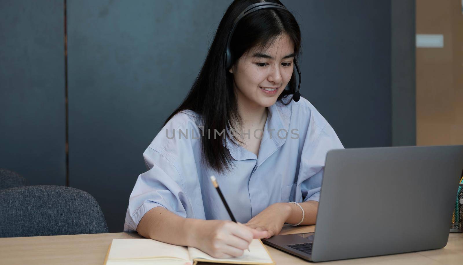Smiling Asian young female using headset looking at laptop screen listen and learning online courses. Happy chinese business woman with headphones video call for customer service by wichayada