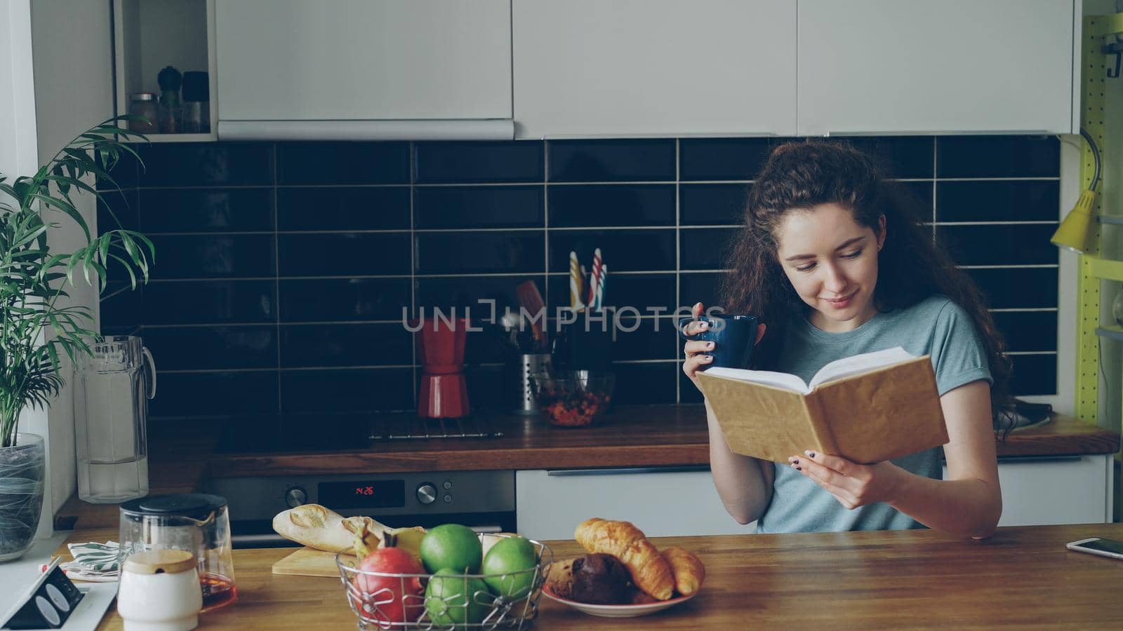 Beautiful curly woman sitting at table holding cup of coffee, drinking and reading book in nice kitchen at home, she is positive and smiling by silverkblack
