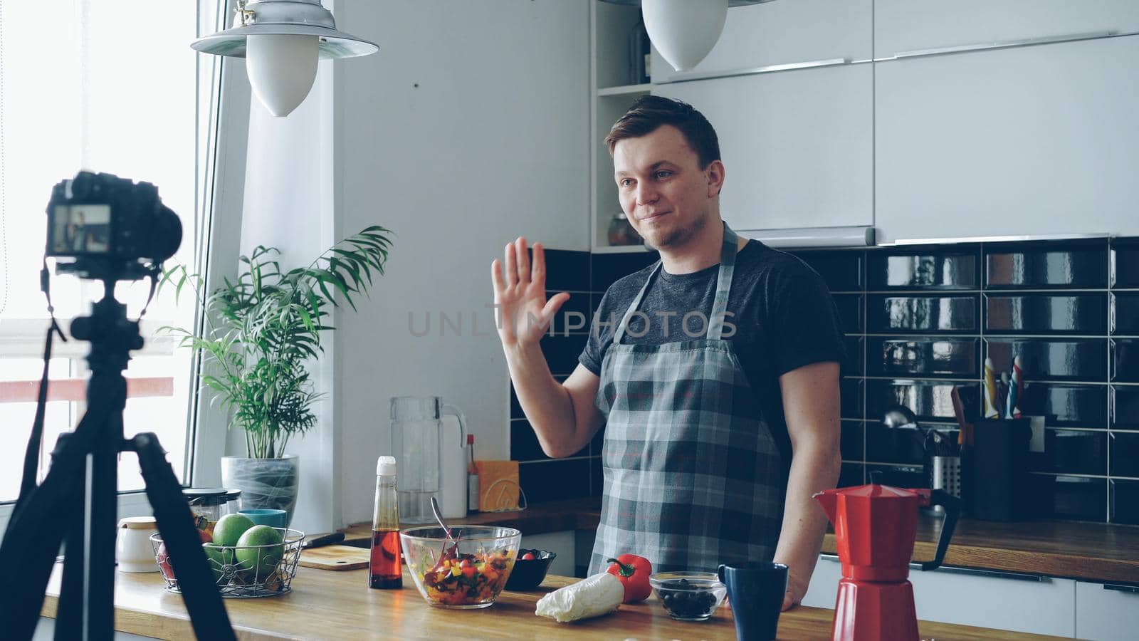 Cheerful attractive man recording video food vlog about healthy cooking on digital camera in the kitchen at home. Vlogging, youth culture and social media concept