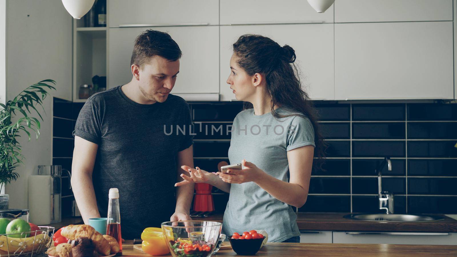 young curly woman showing something unpleasant in husband's phone while he is cooking , they are shouting and quarrelling, man is angry and irritated by silverkblack